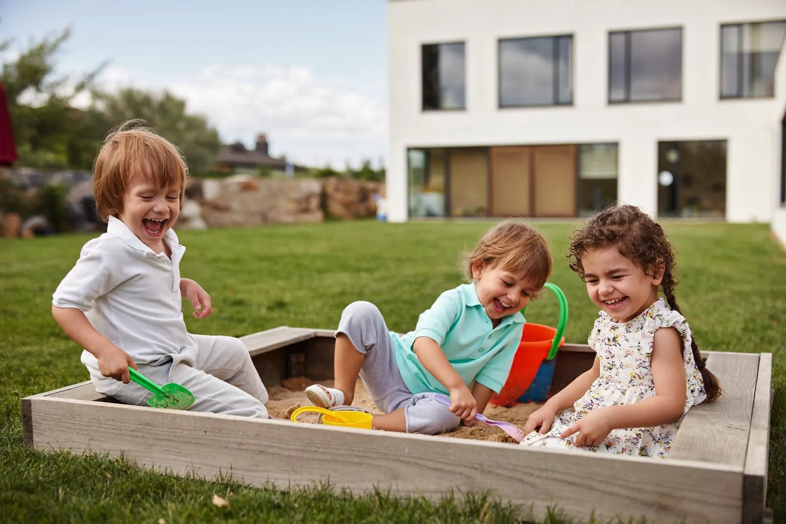Drei Kinder spielen fröhlich im Sandkasten in einem sonnigen Garten